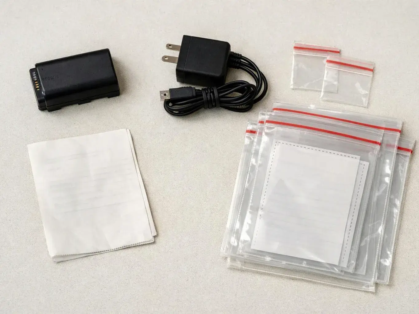 A black battery, charger cord, small plastic bags, and folded white papers lie on a light tabletop