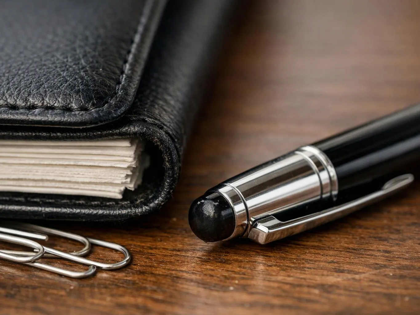 A black leather notebook, silver paper clips, and a black stylus pen rest on a wooden table