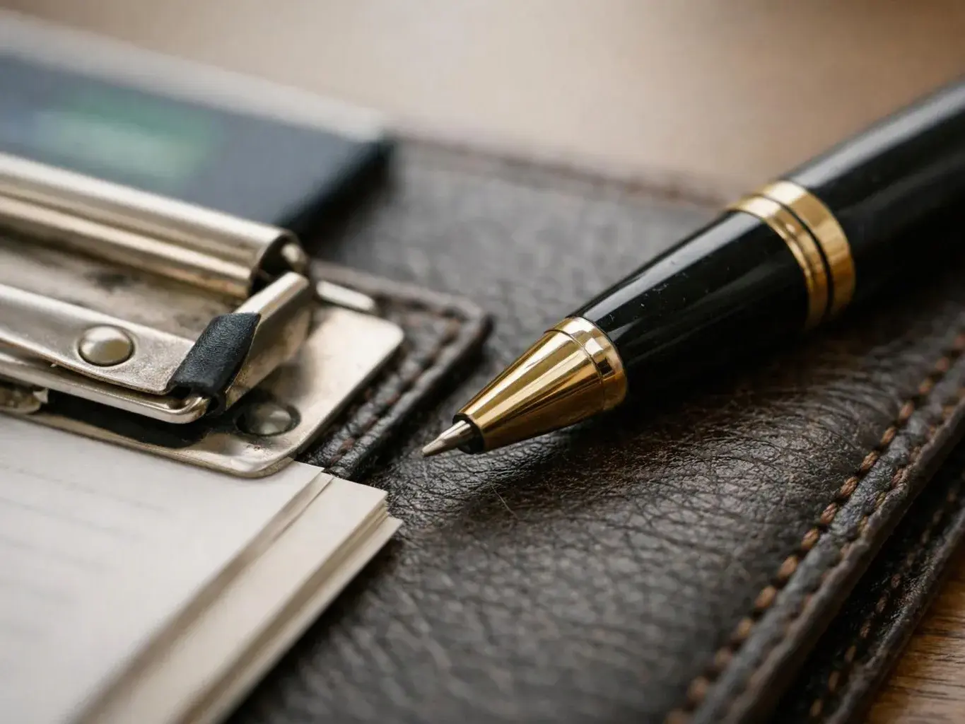 A black pen and a metal clipboard rest on a brown leather desk pad with paper underneath