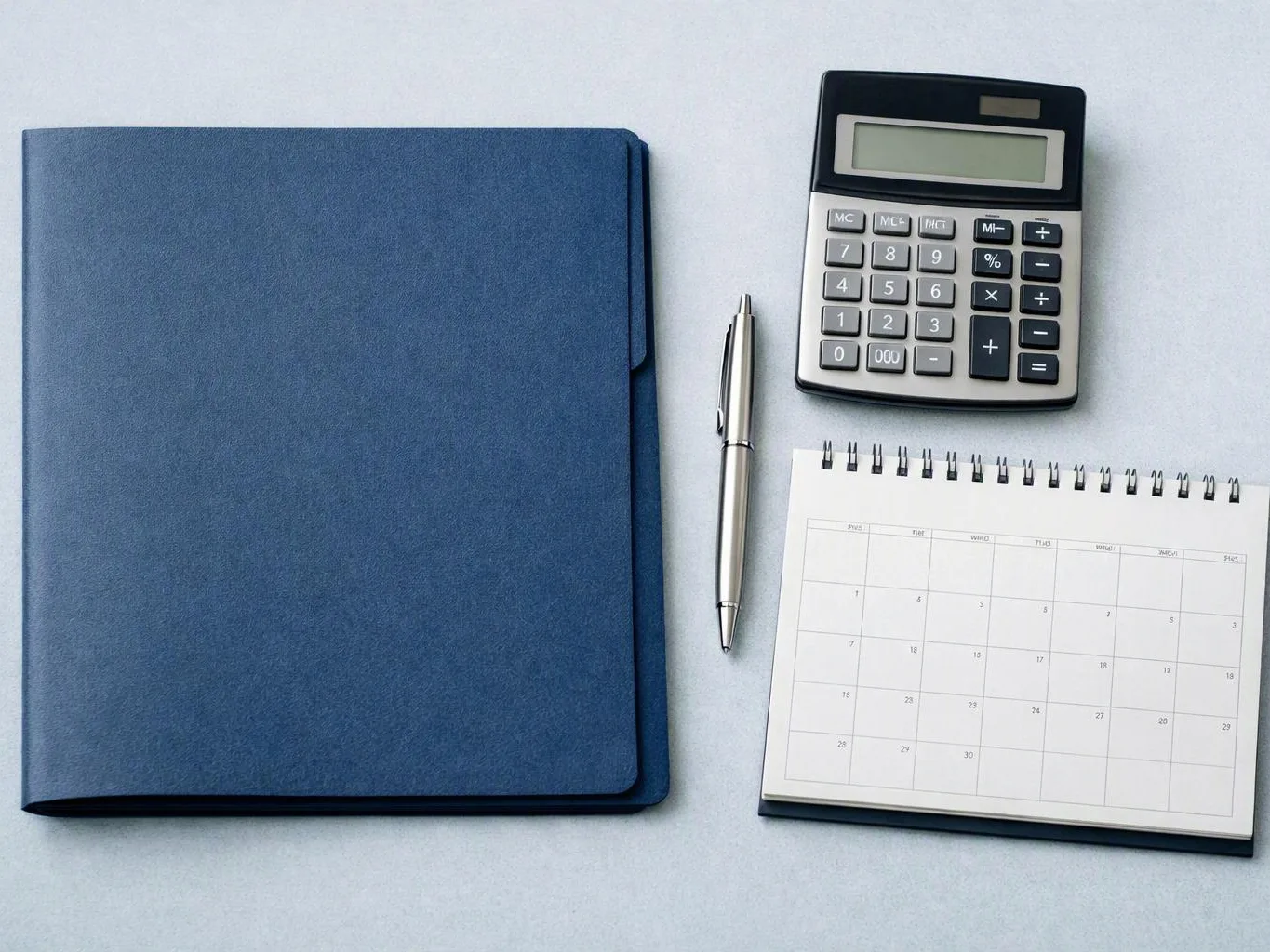 A blue folder, silver pen, calculator, and spiral calendar sit neatly on a light desk surface