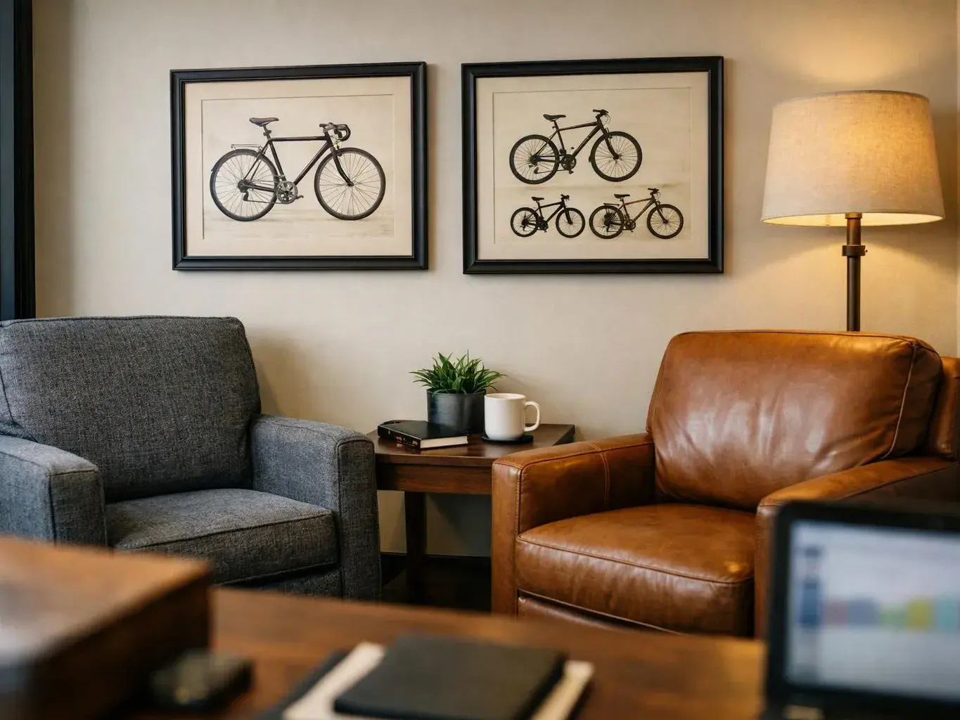 A cozy seating area has two armchairs, framed bicycle prints, a lamp, and a small table with a plant and mug