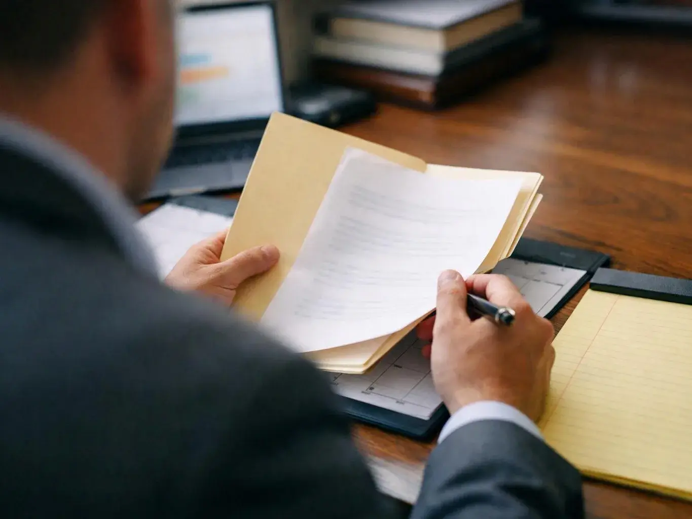 A person in a suit flips through papers at a wooden desk with a laptop and notebooks nearby