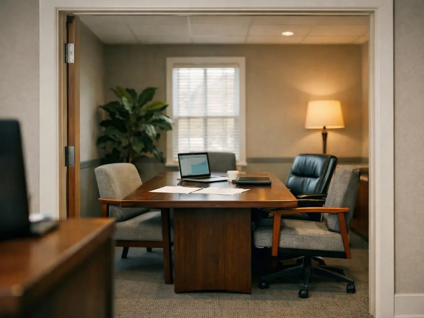 A quiet meeting room holds a wood table, chairs, a laptop, papers, a lamp, and a tall plant