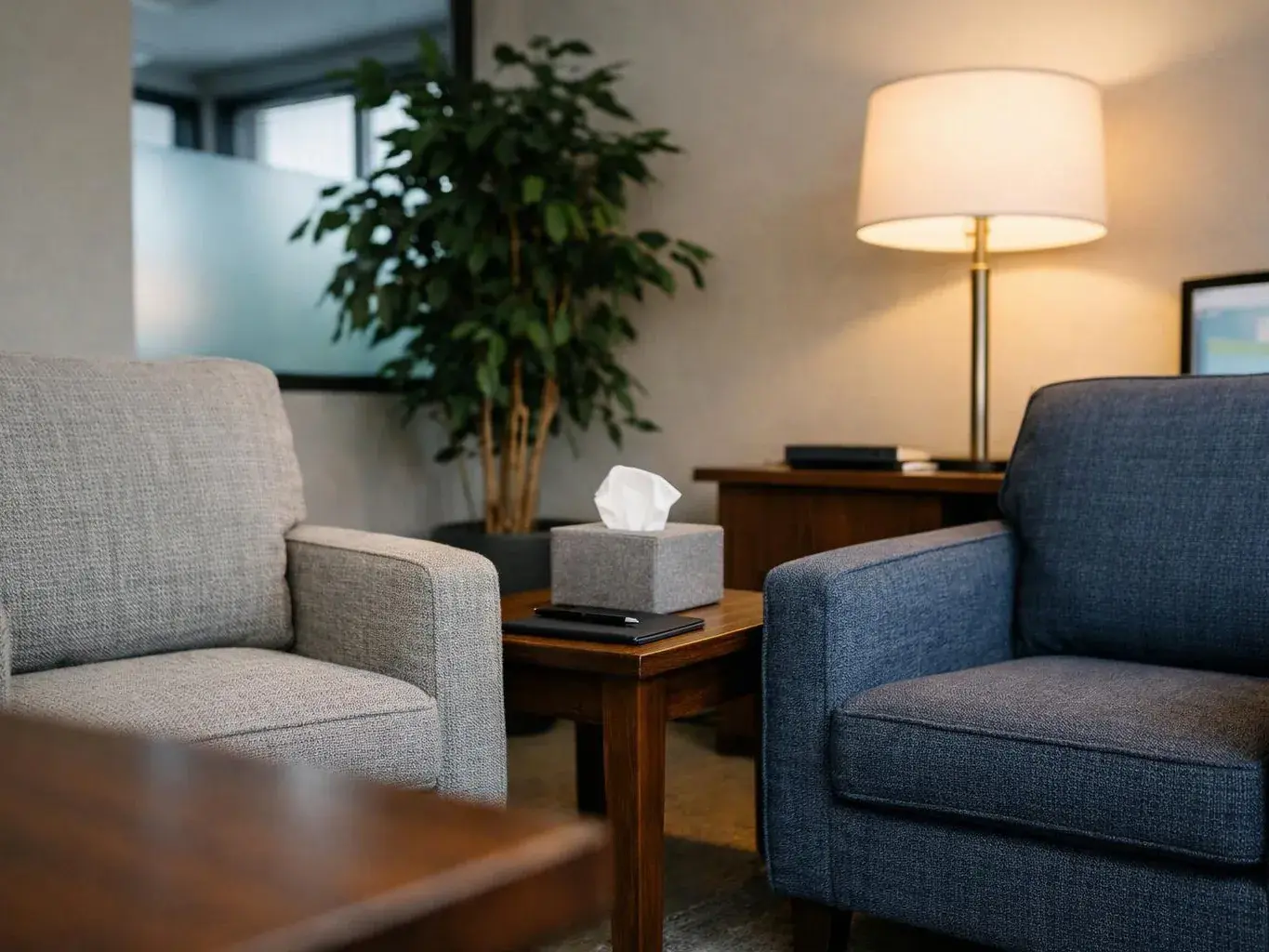 Two empty armchairs face a small table with tissues in a softly lit waiting room